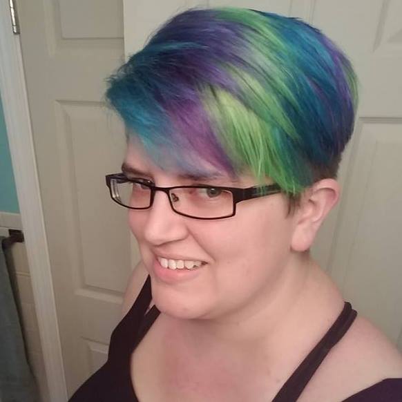 White person in black tank top and rectangular black plastic rimmed glasses looking to camera's left and smiling. Their short hair falls in waves of blue, purple, and green.